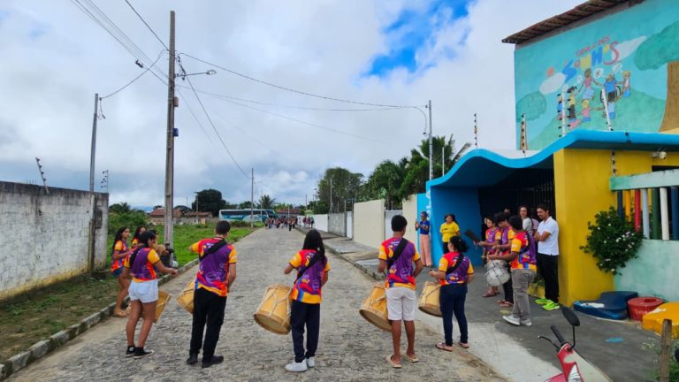 Jovens de costas na rua e na calçada, em frente a uma escola, tocando instrumentos de percussão.