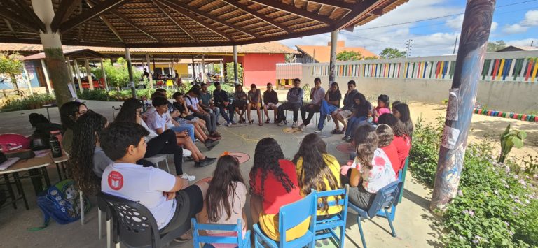 Estudantes reunidos em um pátio, sob um grande gazebo, sentados em cadeiras formando um círculo